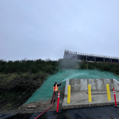 Worker spraying green hydroseed on hillside for erosion control near construction site.