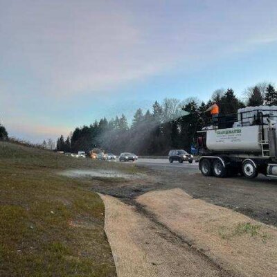 Water truck spraying roadside near highway with passing cars under a hazy sky.
