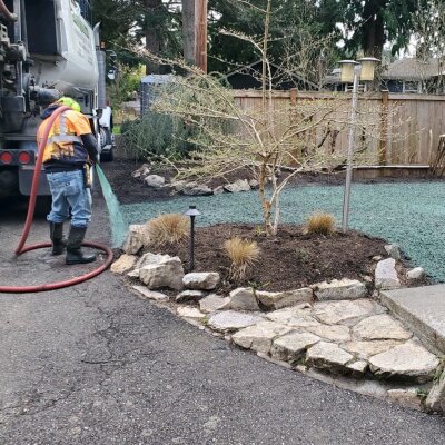 Worker with hose near truck in residential garden with stone path.