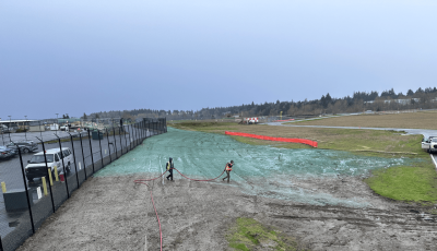 Workers spraying liquid on grass near parking lot and racetrack.