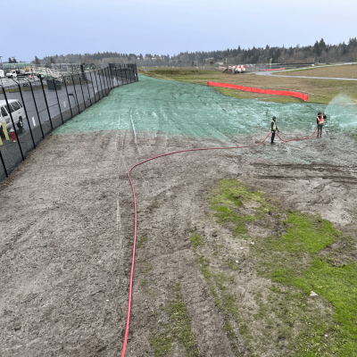 Workers spraying green hydroseed on soil for grass growth near airport.