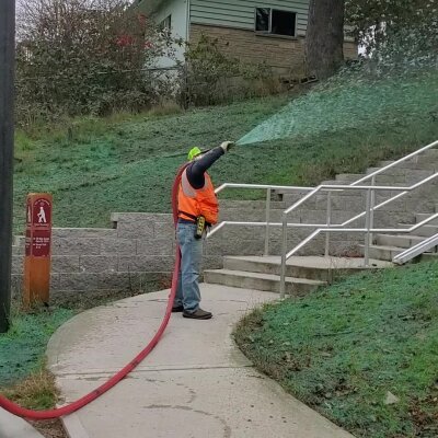 Worker spraying green hydroseed mixture on hillside for erosion control.