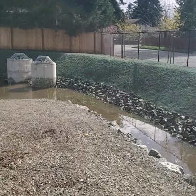 Industrial air vents near a small creek with rocks and chain-link fence in background.