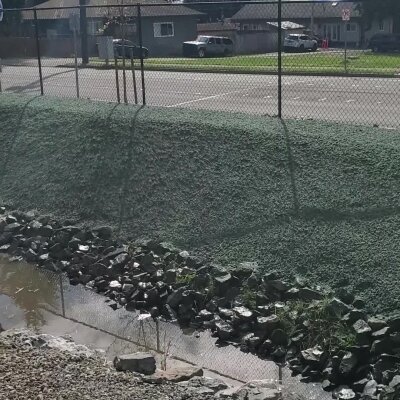 Chain-link fence overlooking water with rocks and green slope.