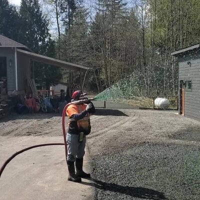 Person using leaf blower on gravel driveway near greenery.