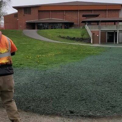 Person in reflective vest standing near grassy area outside a brick building.