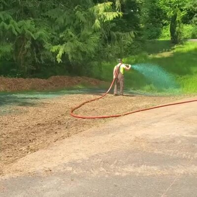 Worker spraying blue-green hydroseed mixture on soil near wooded area in Washington state.